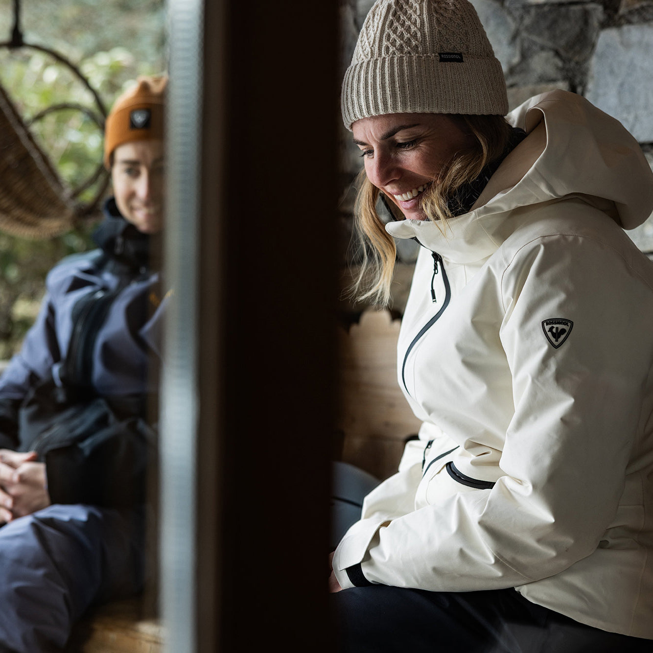 Two people sitting indoors wearing winter jackets and hats.