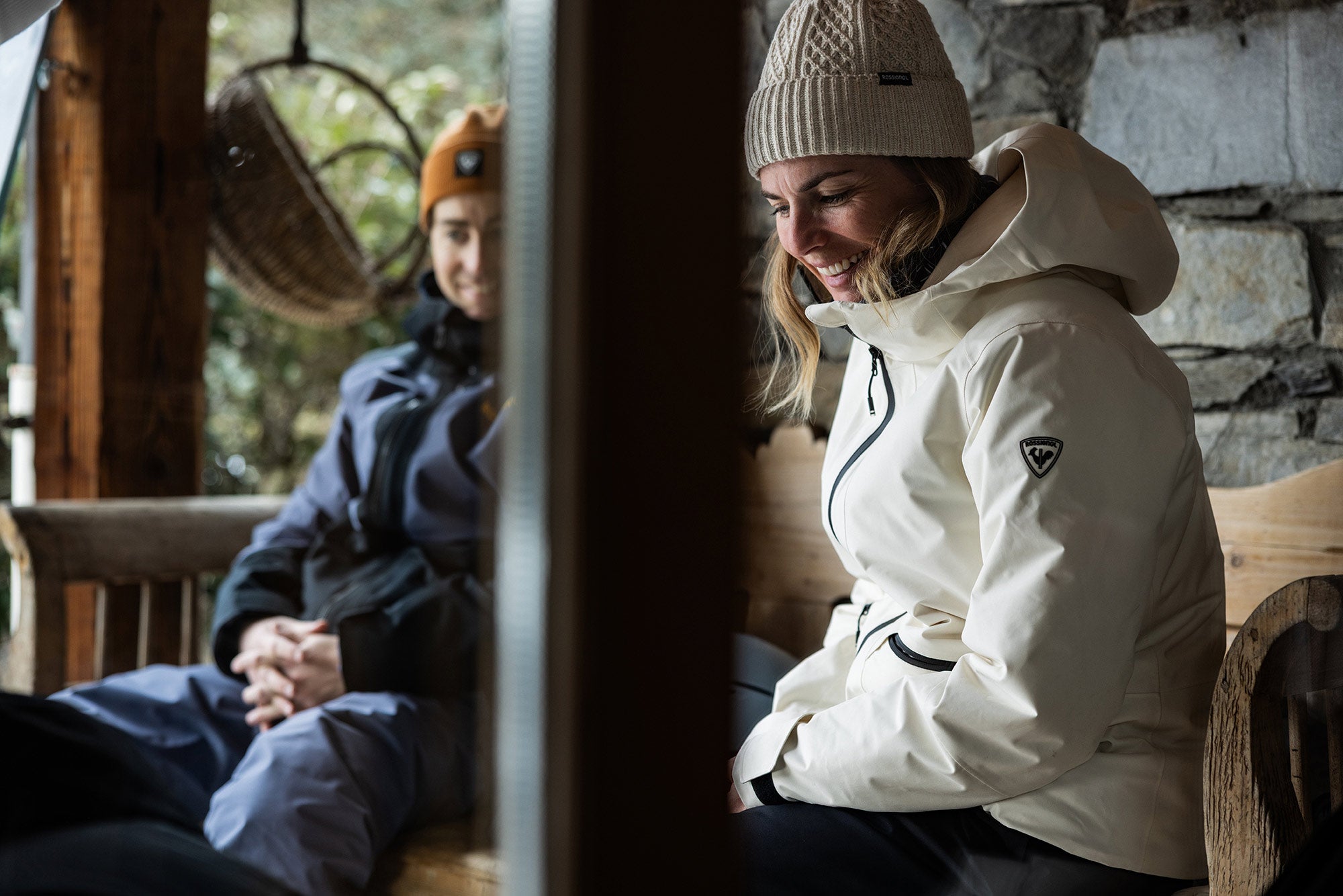 Two people sitting indoors wearing winter jackets and hats.